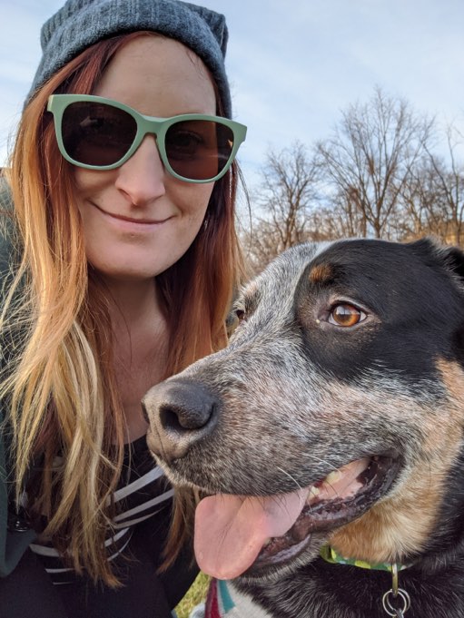 Woman wearing a knit cap and sunglasses crouched next to her blue heeler dog, both of them smiling